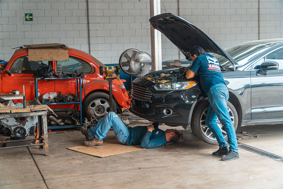 Dos mecánicos trabajando en un taller luminoso, uno bajo un coche y otro en el motor